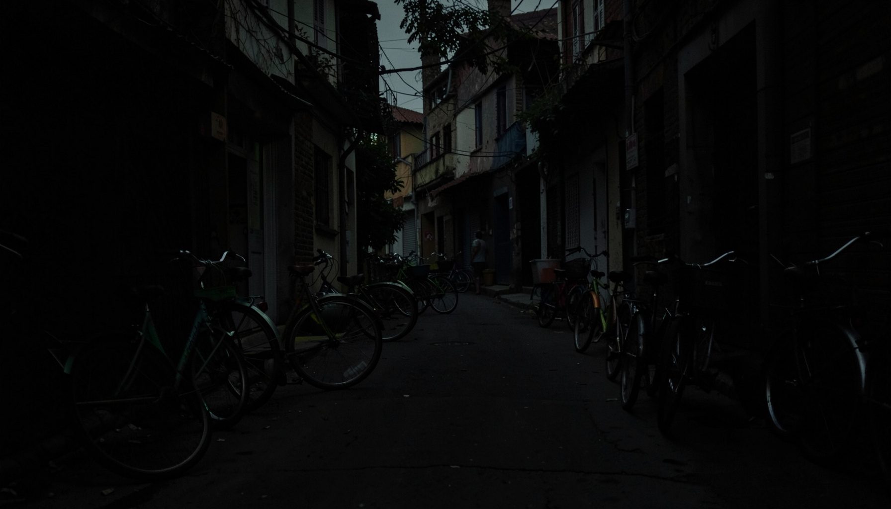 A dark, low-key photograph of a modest neighborhood street with parked tricycles, deep shadows, early evening