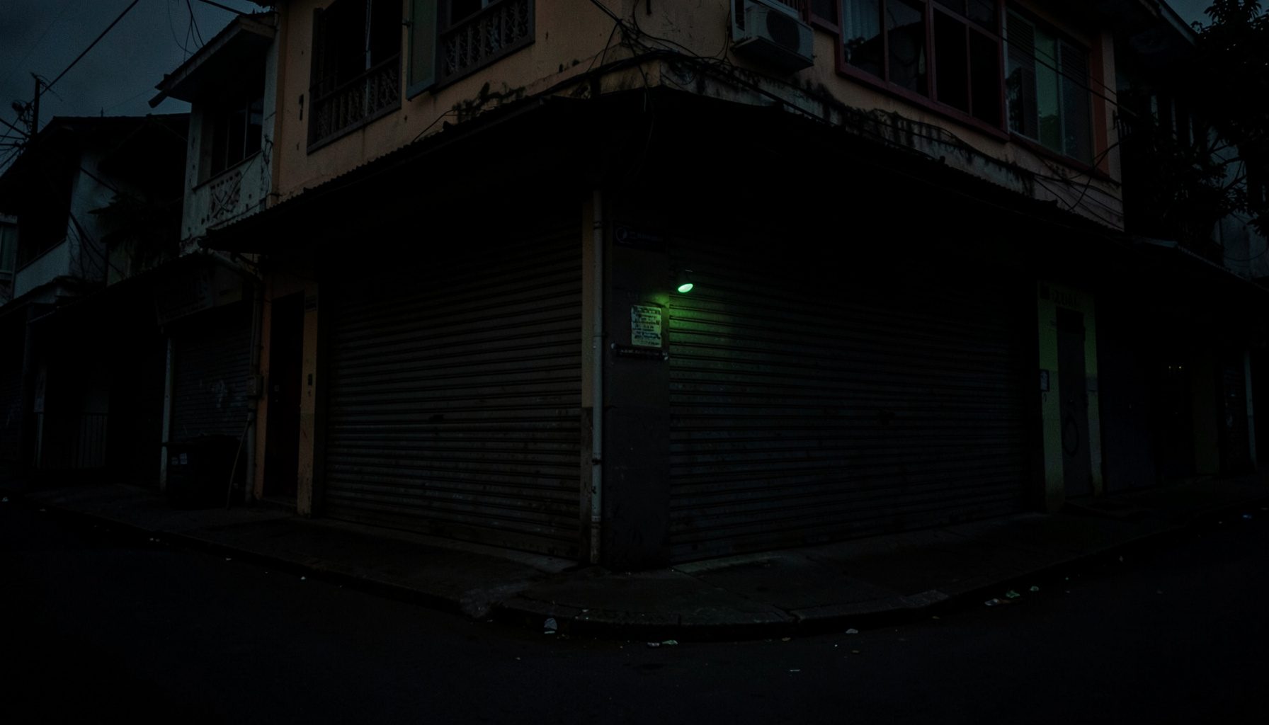 A dark, low-key photograph of an urban street corner in the Philippines at dusk, storefronts with closed shutters, moody atmosphere