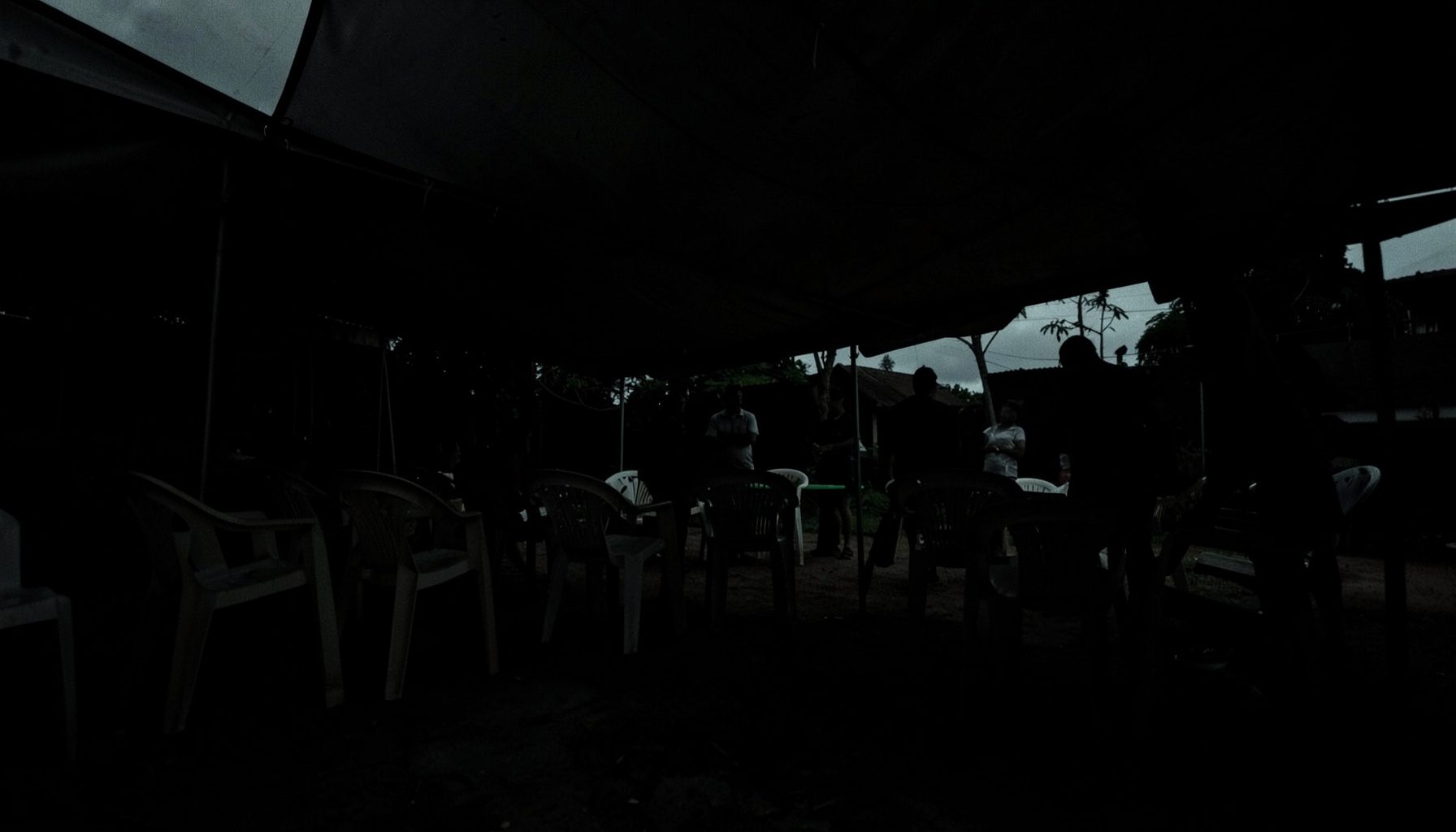 A dark, low-key photograph of a simple outdoor gathering space with plastic chairs arranged under a tent, overcast sky