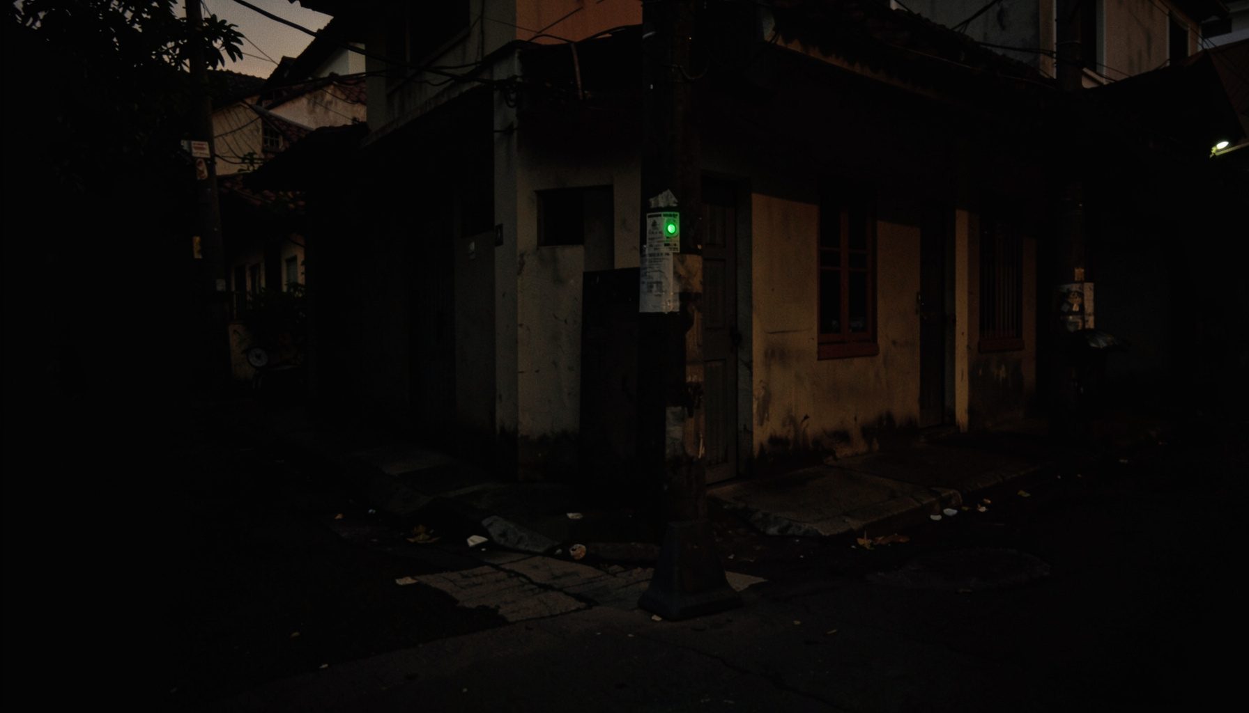 A dark, low-key photograph of a quiet street corner in a Philippine neighborhood at dusk, deep shadows and muted warm tones